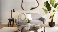 Ruang tamu berdesain minimalis dengan pencahayaan alami. Terdapat sofa abu-abu muda dengan dua bantal di tengah, tepat di bawah cermin dinding asimetris berbingkai kayu. Di sebelah kiri sofa terdapat meja samping hitam dengan lampu baca, dan di sebelah kanan terdapat tanaman hias besar berdaun lebar. Di area depan, terdapat meja kopi kaca bundar dengan tanaman sukulen kecil di atasnya, berpijak pada karpet berwarna terang.